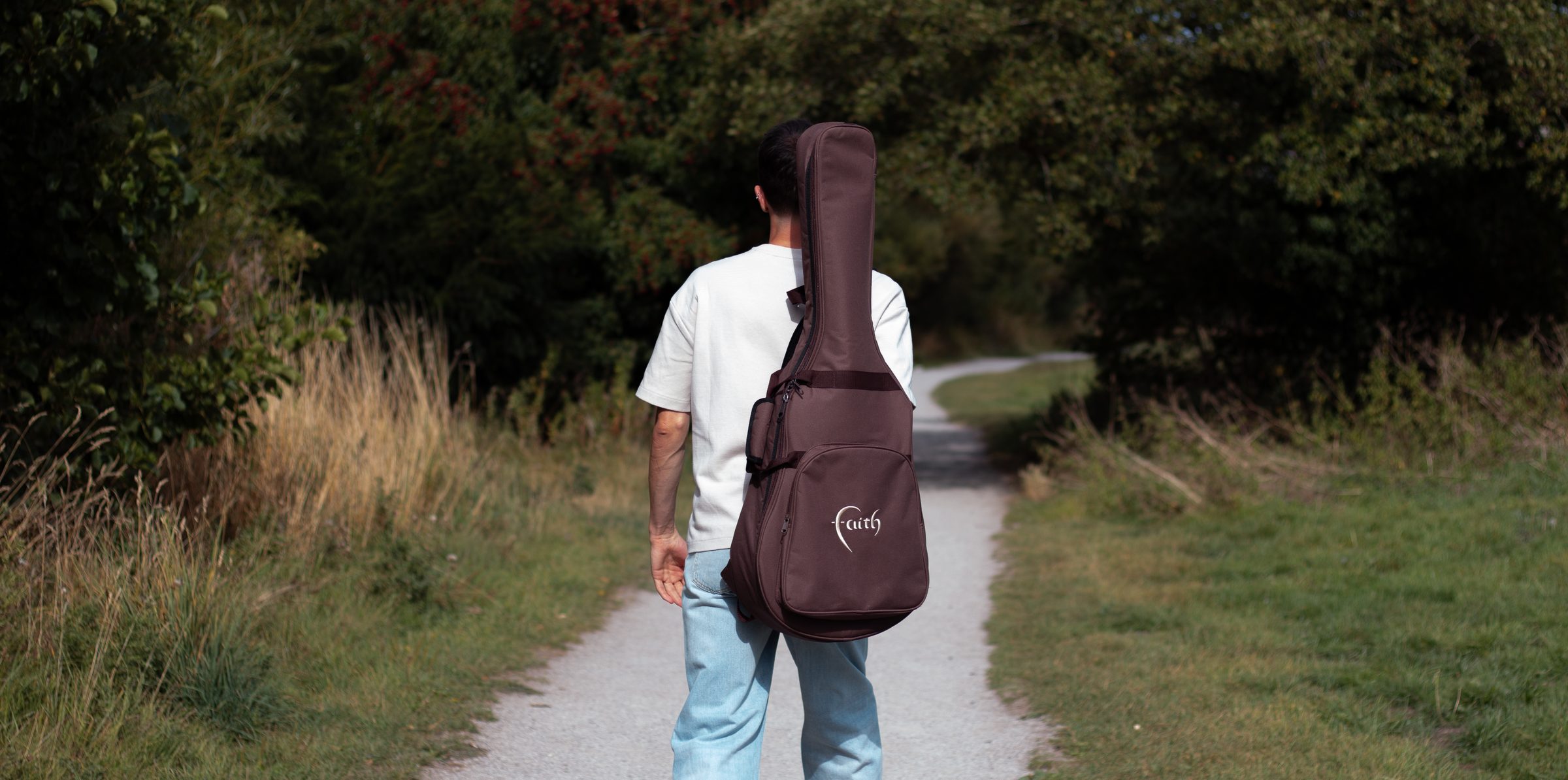 Ben Stafford walking away along a gravel path between summer hedgerows, an acoustic guitar in a soft case slung over his shoulder.